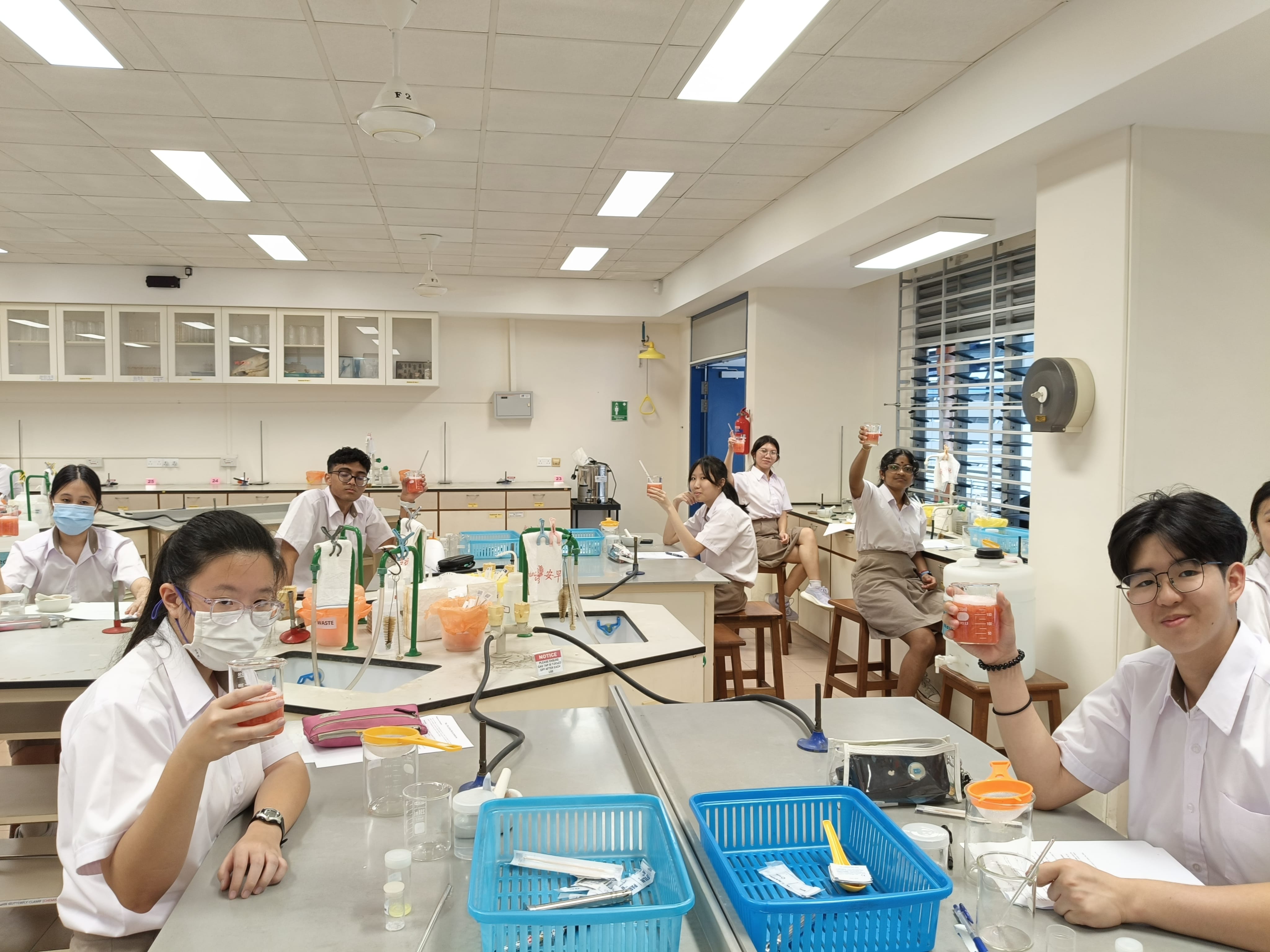 About 10 students in white school uniforms sit at lab benches in a science laboratory, cheerfully holding up beakers of pink-orange liquid from their strawberry DNA extraction experiment.